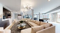 This single-floor amenity space is thoughtfully designed to offer a variety of distinct zones within one cohesive, open layout. The entrance is anchored by a round cream table beneath a brass candelabra chandelier, flanked by sleek black steel-framed partition screens that define intimate seating areas beyond. These partitioned alcoves feature dark upholstered armchairs, curated décor, and pendant lighting, creating a semi-private lounge feel within the broader open plan.

The main lounge area is expansive, outfitted with generous neutral sectional sofas, glass-topped coffee tables, and sculptural sputnik-style chandeliers overhead. A wall-mounted television and foosball table add a recreational dimension to this zone. Light wood flooring runs throughout, complementing the soft grey walls and abundant natural light drawn in by oversized windows overlooking the surrounding building complex.

A communal kitchen serves as a bold focal point, featuring teal cabinetry with brass hardware, dark countertops, and a dramatic floor-to-ceiling marble slab backsplash. Adjacent café-style seating with bistro tables provides a casual dining area. A glazed door opens directly to an outdoor terrace, further enhancing the connection between interior and exterior amenity spaces.