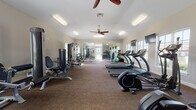 This spacious fitness center is designed to support a full range of workout routines, offering a thoughtfully organized layout across a single open floor. The cardio zone is lined with multiple SportsArt treadmills and elliptical machines positioned along large windows that flood the space with natural light and offer views of the surrounding exterior. Ceiling fans overhead provide comfortable air circulation throughout the workout area.

The strength-training section features a variety of cable machines and multi-function weight stations, complemented by a dedicated dumbbell rack with a full set of free weights. A functional trainer cable system adds versatility for a wide range of exercises. An adjustable weight bench and a collection of medicine balls and stability balls in various sizes round out the functional fitness offerings.

Full-length mirrors line a portion of the walls, enhancing the sense of openness while allowing users to monitor their form during exercise. Wall-mounted televisions are positioned for easy viewing from the cardio equipment. The flooring throughout is carpeted in a neutral tone, and flush and fluorescent lighting fixtures maintain consistent illumination. A clearly marked emergency exit door is accessible from within the space, and overall the facility is clean, bright, and well-maintained.