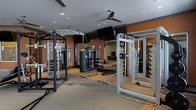 This fitness center is thoughtfully designed to accommodate a wide range of workout styles and training needs. The space features a durable rubberized floor, providing both safety and comfort during exercise. Warm, earthy orange accent walls contrast with a neutral grey ceiling, creating an energizing yet focused atmosphere. Ceiling fans ensure consistent air circulation throughout the space.

The strength training area is well-appointed with a cable crossover machine, a Smith machine loaded with weight plates, and a heavy punching bag suspended from the ceiling — catering to both traditional lifting and functional fitness routines. Weight storage racks and benches are conveniently positioned throughout the floor, supporting efficient transitions between exercises. Full-length mirrors line the walls, enhancing the sense of openness while allowing users to monitor their form during workouts.

Recessed lighting provides bright, even illumination across the entire space, complemented by a wall-mounted flat-screen television. A glass-paneled door or partition adds a modern architectural touch, contributing to the clean, professional aesthetic. Overall, this fitness center offers a comprehensive, well-organized environment suitable for a variety of fitness routines, from strength training to cardio and combat sports practice.