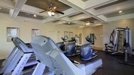 This fitness center is a well-appointed, single-floor facility designed to support a comprehensive workout experience. The space is anchored by a striking coffered ceiling with decorative grid molding, adding an architectural elegance uncommon in typical gym environments. Two ceiling fans are mounted within the coffered panels, ensuring consistent air circulation throughout the room and enhancing comfort during workouts. Recessed lighting supplements the ambient light, keeping the space bright and inviting. The fitness floor is lined with a dark rubber surface, providing durability and traction underfoot. A row of Matrix-brand treadmills runs along one side of the room, complemented by multiple elliptical cross-trainers positioned toward the center and rear of the space. Informational posters and instructional charts are displayed on the walls, offering guidance for users. French-style glass-paned doors are visible along the back wall, adding a refined architectural detail and suggesting additional access points or adjoining areas. The overall ambiance strikes a balance between functional athleticism and polished design, making it a welcoming and motivating environment for fitness enthusiasts of all levels.