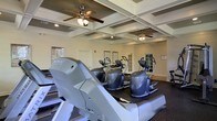 This fitness center is a well-appointed, single-floor facility designed to support a comprehensive workout experience. The space is anchored by a striking coffered ceiling with decorative grid molding, adding an architectural elegance uncommon in typical gym environments. Two ceiling fans are mounted within the coffered panels, ensuring consistent air circulation throughout the room and enhancing comfort during workouts. Recessed lighting supplements the ambient light, keeping the space bright and inviting. The fitness floor is lined with a dark rubber surface, providing durability and traction underfoot. A row of Matrix-brand treadmills runs along one side of the room, complemented by multiple elliptical cross-trainers positioned toward the center and rear of the space. Informational posters and instructional charts are displayed on the walls, offering guidance for users. French-style glass-paned doors are visible along the back wall, adding a refined architectural detail and suggesting additional access points or adjoining areas. The overall ambiance strikes a balance between functional athleticism and polished design, making it a welcoming and motivating environment for fitness enthusiasts of all levels.