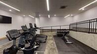 Located at Bridgeport Way W in University Place, Washington, this single-floor fitness facility offers a well-equipped, professionally designed space suited for a variety of workout routines. The layout is organized around a spacious main exercise floor that features a split-level design, with a raised section accessible via a central staircase and bordered by a black metal railing, adding both visual interest and defined workout zones.

The main fitness area is outfitted with a range of cardio equipment, including treadmills, ellipticals, and a stationary cycle, as well as strength-training stations, cable machines, free weights, and medicine ball storage. Flooring throughout the workout areas consists of patterned commercial carpet tiles, while the corridor and transition zones feature large-format gray tile, creating a clear distinction between functional areas.

Linear LED lighting runs along the drop-tile ceiling, providing consistent, bright illumination across the space. Large frosted-glass windows allow diffused natural light into the corridor and ancillary areas without compromising privacy. The walls are finished in crisp white, accented by warm wood-tone baseboards. A water fountain station is conveniently located in the main hallway, alongside clearly marked, ADA-accessible restrooms. The overall aesthetic is clean, functional, and modern.