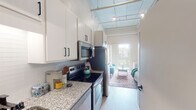 Located on East Lake Street in Minneapolis, this single-floor unit offers a thoughtfully designed open-concept layout that maximizes every inch of its footprint. The kitchen is outfitted with stainless steel appliances, including a range, microwave, and refrigerator, complemented by granite countertops, a subway tile backsplash, and crisp white cabinetry with matte black hardware. An in-unit washer is conveniently integrated into the kitchen area. Light wood-tone flooring runs throughout, lending warmth and visual continuity across the space. Soaring ceilings with exposed industrial elements—including overhead piping and a grid-style ceiling structure—add architectural character and an airy, loft-like feel. The living area flows seamlessly toward a large glass door that opens onto a private balcony, flooding the interior with natural light and offering views of the surrounding brick exterior facade. A dedicated sleeping alcove provides a defined sense of separation within the open plan. The bathroom features a full tub-shower combination with a curved curtain rod and clean neutral finishes. The unit's overall aesthetic blends modern updates with industrial-style design details, creating a functional and stylish urban living space in the heart of Minneapolis.