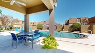 This outdoor recreational space centers around a generously sized freeform swimming pool with clear blue water, framed by a classic brick coping border. The pool area is enclosed within a brick perimeter wall that provides privacy while offering views of the surrounding multi-story brick buildings, mature shade trees, and well-maintained landscaping with trimmed hedges and potted plants.

A covered patio structure with sturdy concrete columns adjoins one side of the pool, providing shaded relief and furnished with a round metal table and matching blue sling chairs — ideal for outdoor dining or relaxing out of direct sun. Adjacent to the covered area, a broad open-air sun deck offers ample room for lounging, outfitted with multiple blue reclining chaise lounges positioned to take full advantage of the abundant natural sunlight.

A poolside amenity building with large windows and a glass-paneled door serves as a support facility for the pool area. The surrounding grounds are further enhanced by mature deciduous trees that provide natural shade and greenery, complementing the warm earth-toned brick aesthetic of the architecture. The entire space is designed for comfortable outdoor recreation under open skies.