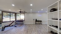 This single-floor fitness facility is thoughtfully designed to support a comprehensive workout experience across multiple functional zones. The space features light-toned luxury vinyl plank flooring throughout, lending a clean, modern aesthetic that ties the entire layout together cohesively.

The cardio zone is bathed in natural light from large corner windows and a glass-paneled exterior door, offering pleasant outdoor views while exercising. Two treadmills, an elliptical, and a stationary bike are positioned to take full advantage of this bright, airy section. A ceiling fan overhead ensures comfortable airflow during high-intensity sessions.

The strength training area is anchored by a full dumbbell rack, an adjustable weight bench, and a cable machine, with floor-to-ceiling mirrors along the wall to assist with form and visually expand the space. Recessed lighting and an additional ceiling fan keep this zone well-lit and well-ventilated.

A separate open area provides ample room for functional training or stretching, accented by a motivational neon sign on the wall. A utility station with a small table and equipment accessories adds practicality to the space. An additional recreational area features a foosball table, a large chalkboard wall, and a mounted display screen, creating a versatile environment suited for both training and community engagement.