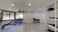 This single-floor fitness facility is thoughtfully designed to support a comprehensive workout experience across multiple functional zones. The space features light-toned luxury vinyl plank flooring throughout, lending a clean, modern aesthetic that ties the entire layout together cohesively.

The cardio zone is bathed in natural light from large corner windows and a glass-paneled exterior door, offering pleasant outdoor views while exercising. Two treadmills, an elliptical, and a stationary bike are positioned to take full advantage of this bright, airy section. A ceiling fan overhead ensures comfortable airflow during high-intensity sessions.

The strength training area is anchored by a full dumbbell rack, an adjustable weight bench, and a cable machine, with floor-to-ceiling mirrors along the wall to assist with form and visually expand the space. Recessed lighting and an additional ceiling fan keep this zone well-lit and well-ventilated.

A separate open area provides ample room for functional training or stretching, accented by a motivational neon sign on the wall. A utility station with a small table and equipment accessories adds practicality to the space. An additional recreational area features a foosball table, a large chalkboard wall, and a mounted display screen, creating a versatile environment suited for both training and community engagement.