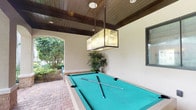 This single-level space is centered around a well-appointed covered outdoor entertainment area designed for leisure and recreation. The focal point of the space is a full-size pool table with a vibrant teal felt surface, positioned beneath a striking geometric pendant light fixture that adds a refined, artistic touch to the setting. The ceiling is finished with warm-toned, rich wood planking accented by dark wood beams, lending a sophisticated yet relaxed ambiance to the outdoor area. The flooring transitions from brick pavers at the perimeter to the covered structure, reinforcing the seamless indoor-outdoor design. A graceful arched opening frames views of the lush surrounding landscaping, allowing natural light to filter in and connecting the covered area to the verdant garden beyond. Tropical potted plants are positioned along the walls, enhancing the organic, resort-like atmosphere. A framed window on the adjacent wall provides an additional architectural detail and a visual connection to the interior. The overall design balances comfort and functionality, making this space well-suited for entertaining guests in a relaxed, outdoor setting enriched by natural greenery and thoughtful architectural detailing.