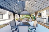 Located at 2220 E 7th St in Charlotte's vibrant Plaza Midwood neighborhood, this outdoor space features a well-appointed covered gazebo patio designed for communal gathering and al fresco dining. The gazebo is anchored by a steeply pitched metal roof with a classic ridge design, strung with globe pendant lights that provide warm ambient illumination for evening use. Multiple slatted dining tables with cushioned swivel chairs offer generous seating capacity beneath the structure, while fabric drape curtains line the perimeter, adding a sense of privacy and style. The concrete patio extends beyond the gazebo's footprint, providing additional open-air space. Surrounding landscaping includes mulched planting beds, decorative stone borders, and manicured shrubbery that frame the area with greenery. A decorative lattice-block retaining wall defines one edge of the courtyard, complementing the contemporary multi-story building façade featuring dark metal balconies, large windows, and a mix of painted brick and panel cladding. Mature trees provide natural shade and contribute to the serene, park-like atmosphere of this thoughtfully designed outdoor amenity space.