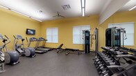 Located at 9393 Tidwell Rd in Houston, Texas, this fitness center is a comprehensively equipped, single-floor exercise facility designed to support a full range of workout routines. The space features warm yellow accent walls paired with neutral tones, rubber flooring throughout for safety and durability, and bright overhead fluorescent lighting that ensures excellent visibility across the entire room. Ceiling fans provide additional air circulation, contributing to a comfortable workout environment.

The cardio section is well-stocked with multiple treadmills, elliptical machines, and a stationary bike, with several wall-mounted flat-screen televisions positioned for easy viewing during exercise. A dedicated strength training area includes a multi-function cable machine, a weight bench, and a comprehensive dumbbell rack offering a wide range of free weights. A full-length wall mirror visually expands the space and allows users to monitor their form. An additional rack holds a variety of medicine balls and exercise balls, supporting functional and cross-training workouts.

Natural light filters in through multiple windows fitted with roller shades, adding brightness while allowing light control. The layout is open and thoughtfully organized, providing ample floor space for movement between equipment zones. This fitness center offers a practical and fully equipped environment for a diverse range of exercise needs.