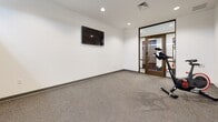 Located in Wichita, KS, this single-floor fitness space offers a well-designed environment tailored for focused exercise. The room features a durable speckled rubber-style floor that provides both traction and cushioning, making it practical for a variety of workout activities. Crisp white walls and recessed ceiling lighting give the space a clean, contemporary aesthetic throughout.

The main workout area is equipped with a Spirit spin bike and wall-mounted fitness accessories, including resistance bands and a set of hanging sandbags, offering versatility for strength and cardio training. A wall-mounted flat-screen television is positioned for easy viewing during workouts. High clerestory windows along one wall allow natural light to filter in while maintaining privacy, contributing to an open and energizing atmosphere.

A glass-paneled door with a warm wood frame separates the fitness area from an adjacent space, adding architectural interest while maintaining visual connectivity. The overall layout is open and uncluttered, providing ample room for movement. The combination of modern finishes, purposeful equipment placement, and thoughtful design makes this a functional and inviting space for fitness use.