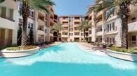 Located at 1901 S Voss Rd in Houston, Texas, this upscale residential community presents a sophisticated architectural design characterized by a warm stucco and stone exterior, large grid-pane windows, and private balconies that overlook a beautifully landscaped interior courtyard. The multi-story building wraps around a stunning resort-style pool as its centerpiece — featuring expansive turquoise waters flanked by lounge chairs, swaying mature palm trees, and meticulously maintained tropical greenery that evokes a vacation-like atmosphere year-round. Residents access the courtyard through decorative wrought-iron gates, with signage indicating dedicated courtyard hours for a well-managed community environment. Adjacent to the pool area, an intimate outdoor seating space offers a patio table and chairs alongside an elegant water fountain feature, ideal for relaxed outdoor gatherings. The building's glass entry doors, framed in sleek metal with recessed lighting above, lend a polished, modern feel to the arrival experience. Brick-paved walkways connect the various outdoor zones, enhancing both the aesthetic and functional flow of the grounds. The overall ambiance of the community is one of refined, resort-inspired living in the heart of Houston.