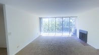 This single-floor space is defined by its clean, open layout and an abundance of natural light. The living area is a standout feature, showcasing a wall of floor-to-ceiling sliding glass doors that flood the interior with sunlight and frame a verdant, tree-lined view. The sliding doors provide direct access to a balcony, seamlessly blending indoor and outdoor living. Neutral-toned carpeting runs throughout the main living area, adding warmth and comfort underfoot, while crisp white walls create a bright, airy ambiance that enhances the sense of openness. The minimalist design allows the natural surroundings and light to take center stage, making the space feel connected to the outdoors. The overall atmosphere is calm and inviting, well-suited for a variety of uses. The combination of generous glazing, outdoor access, and a simple, uncluttered interior gives this space a light-filled, tranquil character that would appeal to those who appreciate natural light and greenery as part of their everyday environment.