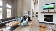 This single-story communal space is defined by its dramatically tall ceilings that lend an open, airy quality throughout. The main lounge area is thoughtfully furnished with a large gray sectional sofa accented by teal and patterned throw pillows, complemented by a tufted gold ottoman and a light blue accent chair. A substantial dark wood multi-drawer dresser serves as a media console, topped with decorative objects and anchored by a large abstract canvas artwork mounted on the wall. Framed black-and-white photography adds a classic touch nearby.

Tall arched windows along one wall flood the space with natural light and offer views of an outdoor pool and surrounding landscaping, with floor-length patterned drapes framing the view elegantly. The flooring throughout is a warm, wood-look plank that ties the various zones together cohesively.

Adjacent to the lounge, a business center nook features a long white shiplap-backed workstation with two computer stations, a shared printer, and industrial-style pendant lighting overhead. The shiplap wall detailing and exposed ceiling beams visible in the lounge add architectural warmth and character, creating a refined yet inviting atmosphere throughout the space.