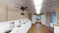 This single-floor laundry facility is designed to accommodate multiple users simultaneously, offering a practical and functional layout. A row of top-loading washing machines lines the left wall, providing ample capacity for residents. Opposite them, a bank of stacked dryer units along the right wall maximizes the use of vertical space, efficiently fitting more machines within the room's footprint.

The flooring throughout is hardwood, lending a cleaner and warmer aesthetic than is typical of utility spaces. Overhead, a combination of fluorescent panel lighting and a ceiling fan with a pull-chain ensures the room remains well-lit and properly ventilated during use. A single window positioned at the far end of the room admits natural light, brightening the space and providing a view of the outdoors.

The walls are painted in a neutral beige tone, contributing to a clean, no-frills environment. An informational board is mounted near the entrance, likely displaying usage instructions or facility guidelines. Overall, the space is straightforward and practical, prioritizing functionality and capacity to serve the needs of a shared residential community.