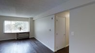 This single-floor space presents a clean, functional layout with a straightforward design. The interior is finished with dark hardwood flooring that contrasts against the bright white walls and white baseboards, creating a crisp, neutral aesthetic throughout. The ceiling features a classic drop tile grid system, which adds a practical, professional quality to the space.

A window fitted with vertical blinds allows natural light to filter into the room, illuminating the open area and highlighting the wall-mounted HVAC unit positioned beneath it — providing both heating and cooling convenience. The walls are kept minimal and unadorned, offering flexibility for various uses or configurations.

A white interior door with a brushed metal lever handle is visible toward one end of the space, suggesting an additional room or closet beyond. A red fire safety device is mounted on the wall, indicating the space meets standard safety requirements. Electrical outlets and switch plates are positioned at regular intervals along the walls.

Overall, the space has a clean, move-in-ready character with neutral finishes that offer a versatile foundation suitable for residential or light commercial use.