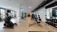 Located at Promenade Vista St in Charleston, SC, this single-floor fitness center is a comprehensively outfitted facility designed to support a wide range of workout routines. The space features light wood-style flooring throughout, white walls, and a clean, modern aesthetic accented by exposed black ductwork that adds an industrial character to the open main floor.

The main strength and training area is expansive, accommodating a broad array of cable machines, multi-functional cable rigs, adjustable benches, weight racks, and free weights. Structural columns are integrated throughout, and ceiling fans maintain airflow across the workout floor. A dedicated cardio zone features Matrix treadmills and stationary bikes positioned in front of large, black-framed windows that overlook palm trees, a pool, and the surrounding community, with wall-mounted televisions providing additional entertainment.

A separate studio space offers cycling bikes, stability balls, BOSU trainers, step platforms, and a full-length mirror wall, making it suitable for group exercise and flexibility training. Another area hosts Matrix cycling bikes alongside kettlebell racks and mirror walls. A punching bag and functional training rig round out the strength training options. The facility's layout supports both individual and group fitness activities in a bright, well-ventilated environment.