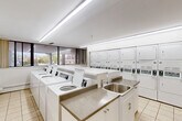 Located at 935 Dundas St E in Mississauga, Ontario, this shared laundry facility offers a clean, functional, and well-organized space designed to serve building residents efficiently. The single-floor layout is thoughtfully divided into distinct zones, each serving a specific purpose.

The main laundry area is the centerpiece of the facility, featuring a row of top-loading washing machines and a bank of stacked dryers along the wall. A stainless steel utility sink with cabinetry below provides a practical hand-washing and pre-treatment station. The space is finished with light ceramic tile flooring throughout and illuminated by bright fluorescent lighting, creating a clean and functional atmosphere. Large windows along the exterior walls bring in natural light and offer views of the surrounding area.

Adjacent to the laundry area is a comfortable waiting room, furnished with padded chairs and side tables, providing residents a pleasant place to wait during laundry cycles. Decorative plants add a touch of warmth to the otherwise utilitarian setting.

A separate multipurpose room features a wall-mounted flat-screen TV, a large mirror along one wall, a folding/work table, and additional large windows overlooking mature trees. Baseboard heating is present throughout, ensuring year-round comfort. Together, these spaces form a practical, resident-friendly amenity facility.