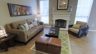 This single-story space centers around a thoughtfully designed living area that balances comfort and style. The room features a striking corner fireplace with a crisp white mantel, serving as a natural focal point and adding warmth to the overall ambiance. Above the mantel, decorative accents complement the space, while framed artwork adorns the surrounding walls, adding color and personality.

The flooring consists of hardwood-style planks in a warm tone, grounding the room with a clean, cohesive look. A patterned area rug in green and white defines the seating area, providing a vibrant contrast to the neutral color palette of the walls and furnishings. The living area is furnished with a generously sized upholstered sofa adorned with decorative pillows, paired with leather ottomans and a dark rectangular coffee table, creating a functional and inviting arrangement.

Natural light enters through large windows fitted with horizontal blinds, brightening the space while maintaining privacy. The overall atmosphere is welcoming and well-coordinated, with a harmonious blend of textures and tones that make the space feel both livable and polished.