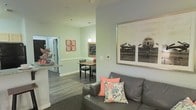 This single-floor residence offers a thoughtfully designed open-concept layout that seamlessly connects the living, dining, and kitchen areas into one cohesive and functional space. The living area features a comfortable gray leather sofa accented with coral and floral throw pillows, anchored by a large framed vintage aviation photograph that serves as a striking focal point on the soft sage-green wall. The space flows naturally into a cozy dining nook, illuminated by a pendant light that casts a warm glow over the compact dining table and chairs. A chalkboard wall feature adds a charming, personalized touch to the dining area, complemented by decorative framed artwork in coral tones. The kitchen is partially open to the living space, offering a convenient pass-through counter that enhances the sense of connectivity throughout the home. Rich, dark hardwood-style flooring runs through the main areas, providing a warm contrast to the light, neutral wall tones. The overall ambiance is bright, inviting, and well-appointed, with a clean and modern aesthetic that is both practical and visually appealing. The thoughtful use of color accents and decorative elements gives the space a personalized, lived-in character.