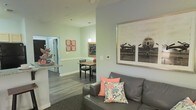 This single-floor residence offers a thoughtfully designed open-concept layout that seamlessly connects the living, dining, and kitchen areas into one cohesive and functional space. The living area features a comfortable gray leather sofa accented with coral and floral throw pillows, anchored by a large framed vintage aviation photograph that serves as a striking focal point on the soft sage-green wall. The space flows naturally into a cozy dining nook, illuminated by a pendant light that casts a warm glow over the compact dining table and chairs. A chalkboard wall feature adds a charming, personalized touch to the dining area, complemented by decorative framed artwork in coral tones. The kitchen is partially open to the living space, offering a convenient pass-through counter that enhances the sense of connectivity throughout the home. Rich, dark hardwood-style flooring runs through the main areas, providing a warm contrast to the light, neutral wall tones. The overall ambiance is bright, inviting, and well-appointed, with a clean and modern aesthetic that is both practical and visually appealing. The thoughtful use of color accents and decorative elements gives the space a personalized, lived-in character.