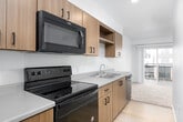 This single-floor residence offers a thoughtfully designed open-concept layout that seamlessly connects the kitchen and living areas. The kitchen is outfitted with warm wood-toned cabinetry featuring matte black hardware, light quartz-style countertops, a stainless steel double sink, and a suite of black appliances including an electric range, over-the-range microwave, and dishwasher. Upper cabinets with open shelving provide both storage and visual interest. The living area is generously proportioned, featuring soft wall-to-wall carpeting and large windows that invite an abundance of natural light. A full-width sliding glass door opens directly to a private balcony, offering views of the surrounding neighborhood and bringing an airy, connected feel to the interior. The bedroom is equally well-lit, with multiple windows and balcony access, continuing the carpeted flooring and neutral color palette. The bathroom presents a clean, modern aesthetic with light hardwood-style flooring, a wood-vanity cabinet with a countertop sink, a large mirror, and crisp white walls. Throughout the residence, the consistent use of warm wood tones, neutral whites, and black accents creates a cohesive, contemporary ambiance well-suited for comfortable everyday living.