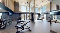 The Modera Gym in Pine Brook, NJ is a thoughtfully designed, expansive fitness facility that delivers a premium workout experience across a single, open floor. The space is defined by soaring double-height ceilings and oversized windows that line the perimeter, flooding the interior with natural light and offering views of the surrounding landscape. Durable rubber flooring spans the entire facility, providing a safe and functional surface throughout. The cardio zone features a row of modern treadmills, stair climbers, and cycling bikes, many positioned directly in front of large windows to enhance the workout atmosphere. A dedicated strength training area is equipped with an extensive dumbbell rack, adjustable benches, cable machines, and a multi-functional cable tower, all supported by full-length mirrors that reinforce the sense of openness. Selectorized weight machines are thoughtfully arranged in a separate section for targeted muscle training. A bold design aesthetic is evident throughout, with dark navy accent walls, decorative wood slat paneling, a textured statement wall, and sleek pendant lighting. Wall-mounted televisions add a modern convenience. The layout flows naturally between zones, making the gym both functional and visually striking.