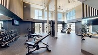 The Modera Gym in Pine Brook, NJ is a thoughtfully designed, expansive fitness facility that delivers a premium workout experience across a single, open floor. The space is defined by soaring double-height ceilings and oversized windows that line the perimeter, flooding the interior with natural light and offering views of the surrounding landscape. Durable rubber flooring spans the entire facility, providing a safe and functional surface throughout. The cardio zone features a row of modern treadmills, stair climbers, and cycling bikes, many positioned directly in front of large windows to enhance the workout atmosphere. A dedicated strength training area is equipped with an extensive dumbbell rack, adjustable benches, cable machines, and a multi-functional cable tower, all supported by full-length mirrors that reinforce the sense of openness. Selectorized weight machines are thoughtfully arranged in a separate section for targeted muscle training. A bold design aesthetic is evident throughout, with dark navy accent walls, decorative wood slat paneling, a textured statement wall, and sleek pendant lighting. Wall-mounted televisions add a modern convenience. The layout flows naturally between zones, making the gym both functional and visually striking.