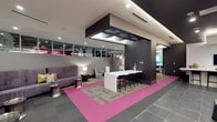 This dynamic single-floor commercial space showcases a striking contemporary design that blends bold color choices with modern architectural details. The interior immediately draws attention with its vivid pink area flooring, which contrasts sharply against the surrounding slate-toned tile and patterned carpet tiles, creating distinct zones within the open-plan layout. A bold, dark suspended ceiling fixture anchors the central area, adding architectural depth and visual interest overhead, complemented by recessed lighting throughout.

The space is thoughtfully divided into functional areas. A sleek white bar-height counter with black barstools serves as a central hub, ideal for collaborative work or informal gatherings. Adjacent to it, a comfortable lounge area features a plush gray sofa, accent chairs, and stylish side tables, providing a relaxed setting for conversation or breakout sessions. The dark accent walls add contrast and definition, while a large display screen is visible in the background, suggesting the space is equipped for presentations or media use.

Expansive interior windows along one wall allow light to flow between areas and maintain visual connectivity throughout the space. The overall ambiance is energetic and professional, reflecting a thoughtfully curated environment well-suited for a modern office, showroom, or collaborative workspace.