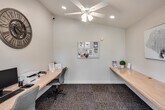 Located at 27550 Hillcrest in Mission Viejo, California, this single-floor business center offers a practical and well-appointed workspace designed to support productivity in a comfortable setting. The room features continuous built-in wooden countertop desks lining multiple walls, providing ample workspace for several users simultaneously. Comfortable task chairs accompany the workstations, making the space well-suited for focused work sessions.

Natural light filters in through windows fitted with horizontal blinds, offering a bright yet controllable environment. A glass-paneled entry door flanked by additional windows further enhances the sense of openness and provides a view of the surrounding greenery outside. The flooring consists of durable, dark-toned carpet tiles that ground the space with a professional feel.

Overhead, a ceiling fan with integrated lighting ensures comfortable air circulation, while recessed lighting provides consistent, even illumination throughout. A cork bulletin board mounted on one wall serves as an information hub, and decorative touches such as framed artwork, a classic gear-style wall clock, and small potted plants add warmth to the otherwise clean, neutral aesthetic. The crisp white walls and uncluttered layout contribute to an organized, focused atmosphere well-suited for a shared community or professional workspace.
