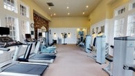 Located at 900 Beverly Hills Rd in Hattiesburg, Mississippi, this fitness center offers a well-appointed, single-floor gym designed to accommodate a comprehensive workout experience. The space features warm yellow walls, recessed lighting, and a generous ceiling height that creates an open, energizing atmosphere. A distinctive stone archway accent wall serves as a charming architectural focal point, adding character to the interior.

The gym is fully equipped with a wide array of cardiovascular machines, including treadmills and a recumbent exercise bike, positioned to allow ample space between units. Along the opposite side, a row of Nautilus strength-training machines provides users with targeted resistance options. A large standing floor fan ensures comfortable air circulation throughout the workout area.

Wall-mounted sconces contribute to the warm ambient lighting, complementing the recessed ceiling lights above. Decorative "GYM" lettering adorns one wall, reinforcing the space's dedicated fitness purpose. A wall clock and informational signage are conveniently placed for easy reference during workouts. The neutral carpeted flooring ties the room together, providing a clean and functional surface throughout. Overall, the space is thoughtfully organized to support both cardio and strength training in a comfortable, well-maintained environment.