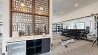 This modern fitness center offers a generously sized, single-floor layout designed to accommodate a full range of workout activities. The space is defined by its crisp white walls, dark baseboard trim, and plush patterned carpet flooring that provides both comfort and durability underfoot. Large black-framed windows line the walls, flooding the interior with abundant natural light and offering open views of the surrounding outdoor greenery, creating an inviting and energizing atmosphere.

The cardio zone is well-appointed with treadmills, elliptical trainers, a stair climber, and stationary bikes, all positioned to take advantage of the garden views. The strength training area features a comprehensive rack of dumbbells, weight benches, and cable machines, complemented by full-length mirrors that enhance the sense of openness. A ceiling fan overhead contributes to comfortable air circulation throughout the space.

A standout design feature is a decorative wooden lattice screen that partitions a small service counter area, complete with a beverage refrigerator and storage cabinetry, adding a warm, textural contrast to the otherwise contemporary aesthetic. Recessed lighting throughout ensures the space remains bright and well-lit at all times. The fitness center is accessed through a glass-paneled entry door, reinforcing the open, transparent design philosophy of the overall space.