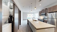 This contemporary single-floor residence showcases a thoughtfully designed open-concept layout that seamlessly connects the kitchen and living areas. The kitchen is a standout feature, centered around a generous island with a pristine quartz countertop, undermount sink, and stylish Edison-bulb pendant lighting. Dark-toned cabinetry pairs elegantly with stainless steel appliances, including a built-in microwave, range, and refrigerator, while light wood-look flooring ties the space together with a warm, modern aesthetic.

Large windows flood the living area with natural light and frame charming views of a lively downtown streetscape, complete with mature trees and historic architecture. A sliding door closet system adds practical storage near the main living space.

The bedroom is carpeted for comfort and features a generous window with outdoor views, creating a bright and peaceful retreat. A hallway connects the bedroom to the bathroom, which is finished with stone-look tile flooring, a dark wood vanity with quartz countertop, a large mirror with overhead lighting, and a bathtub with shower. Throughout the unit, dark-trimmed doors and baseboards provide a refined contrast to the light neutral walls, lending the space a polished, cohesive character.