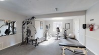 Located at 1601 Hillcrest Road in Mobile, Alabama, this fitness center offers a dedicated, single-floor space designed to support a well-rounded exercise routine. The facility is laid out across multiple connected areas, each serving a distinct workout purpose.

The main exercise room features neutral grey walls, durable carpet flooring, and a textured ceiling with overhead and natural lighting supplied by several windows. Strength training is supported by a multi-station cable machine, a Cybex adjustable bench, and a stability ball, while a reference chart on the wall aids in proper exercise form. Cardio equipment includes an elliptical trainer, a recumbent stationary bike, and a Star Trac treadmill, all positioned to take advantage of available natural light from exterior-facing windows.

The entry and transition area includes safety signage, a fire extinguisher, and clearly posted fitness center rules and guidelines. A hallway leads to additional utility spaces, including what appears to be a restroom or changing area with tile flooring.

The overall ambiance is clean, functional, and well-organized, with a straightforward layout that maximizes usable floor space. The facility is open 24 hours and requires an amenity key for access, making it a convenient option for residents seeking a reliable on-site workout space.