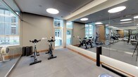 This contemporary fitness center is thoughtfully designed to support a wide range of workout routines within a clean, professional environment. The space is organized into distinct functional zones, separated by sleek floor-to-ceiling glass partitions that maintain an open, airy feel while defining each area.

The main strength and cardio zone features a robust selection of equipment, including a dumbbell rack, cable machines, and cardio machines such as treadmills and ellipticals. Wall-mounted flat-screen televisions are positioned throughout for convenience during workouts. Circular flush-mounted ceiling lights provide consistent, even illumination across the space.

A dedicated studio area accommodates spin bikes and functional training accessories, including stability balls in varying sizes, a BOSU balance trainer, an ab wheel, and a plyometric jump box. This zone benefits from large floor-to-ceiling windows that flood the room with natural light and offer views of an outdoor courtyard, creating an energizing atmosphere.

Throughout the fitness center, large-format gray tile flooring provides a durable and cohesive foundation, complemented by warm wood baseboards and neutral gray walls. Full-length mirrors amplify the sense of space and allow users to monitor their form. The overall design balances functionality with a polished, modern aesthetic suited to a high-quality facility.