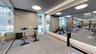 This contemporary fitness center is thoughtfully designed to support a wide range of workout routines within a clean, professional environment. The space is organized into distinct functional zones, separated by sleek floor-to-ceiling glass partitions that maintain an open, airy feel while defining each area.

The main strength and cardio zone features a robust selection of equipment, including a dumbbell rack, cable machines, and cardio machines such as treadmills and ellipticals. Wall-mounted flat-screen televisions are positioned throughout for convenience during workouts. Circular flush-mounted ceiling lights provide consistent, even illumination across the space.

A dedicated studio area accommodates spin bikes and functional training accessories, including stability balls in varying sizes, a BOSU balance trainer, an ab wheel, and a plyometric jump box. This zone benefits from large floor-to-ceiling windows that flood the room with natural light and offer views of an outdoor courtyard, creating an energizing atmosphere.

Throughout the fitness center, large-format gray tile flooring provides a durable and cohesive foundation, complemented by warm wood baseboards and neutral gray walls. Full-length mirrors amplify the sense of space and allow users to monitor their form. The overall design balances functionality with a polished, modern aesthetic suited to a high-quality facility.