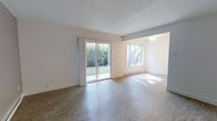 This single-floor space offers a clean, open layout filled with natural light. The main living area is anchored by large sliding glass doors dressed with sheer white curtains, opening directly to a verdant outdoor green space and providing a seamless indoor-outdoor connection. An adjacent picture window further enhances the brightness of the room, allowing sunlight to pour across the smooth, wood-look laminate flooring throughout.

The open-concept design connects the living area to a secondary space through a wide, open archway, creating a fluid and spacious feel ideal for both everyday living and entertaining. A flush ceiling light fixture in the adjoining area adds functional illumination to complement the abundant natural light.

Walls are finished in a crisp white tone, contributing to the airy and clean atmosphere of the space. The textured popcorn ceiling runs throughout, adding a classic residential character. A baseboard heating unit runs along the exterior wall beneath the windows, offering practical climate control.

Overall, this space presents a bright, functional, and well-connected interior with consistent flooring, neutral finishes, and a strong connection to the outdoors through its generous glazing and garden-facing sliding door access.