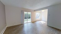 This single-floor space offers a clean, open layout filled with natural light. The main living area is anchored by large sliding glass doors dressed with sheer white curtains, opening directly to a verdant outdoor green space and providing a seamless indoor-outdoor connection. An adjacent picture window further enhances the brightness of the room, allowing sunlight to pour across the smooth, wood-look laminate flooring throughout.

The open-concept design connects the living area to a secondary space through a wide, open archway, creating a fluid and spacious feel ideal for both everyday living and entertaining. A flush ceiling light fixture in the adjoining area adds functional illumination to complement the abundant natural light.

Walls are finished in a crisp white tone, contributing to the airy and clean atmosphere of the space. The textured popcorn ceiling runs throughout, adding a classic residential character. A baseboard heating unit runs along the exterior wall beneath the windows, offering practical climate control.

Overall, this space presents a bright, functional, and well-connected interior with consistent flooring, neutral finishes, and a strong connection to the outdoors through its generous glazing and garden-facing sliding door access.
