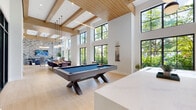 Located at 1711 N University Dr in Plantation, FL, this contemporary amenity space is defined by its dramatic double-height ceilings adorned with warm wood beam detailing, creating an airy and sophisticated atmosphere throughout. Expansive black-framed floor-to-ceiling windows and glass doors flood the interior with natural light while framing lush, verdant views of the surrounding tropical landscaping.

The open-plan layout seamlessly connects several distinct functional areas. A central game area accommodates a full-size billiards table set atop light wide-plank hardwood flooring. Adjacent to this, a lounge area features plush seating arranged around a bold whitewashed brick accent wall, upon which a large flat-screen television is mounted above a sleek media console. Pendant globe lighting adds warmth to this inviting gathering zone.

A dining area with a communal wood table and upholstered chairs is bathed in sunlight streaming through the towering glass walls. The kitchenette is outfitted with quartz countertops, slate-toned cabinetry, a matte black industrial-style faucet, and an integrated sink — all complemented by garden views. Throughout the space, clean white walls, light-toned tile and hardwood floors, and minimalist design choices contribute to a cohesive, modern aesthetic ideal for community gathering and recreation.