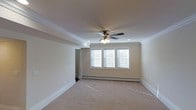 This single-floor space offers a clean and classic interior design with thoughtful architectural details throughout. The main room is generously proportioned, finished with soft, neutral-toned wall-to-wall carpeting that adds warmth and comfort underfoot. Crown molding lines the perimeter of the ceiling, lending an elegant and polished character to the space.

Overhead, a traditional ceiling fan with an integrated light fixture serves as a functional centerpiece, complemented by recessed lighting that provides even, ambient illumination throughout the room. The ceiling itself features a subtle stepped or tray detail, adding visual depth and dimension.

Natural light fills the room through a set of triple windows fitted with white horizontal blinds, allowing for flexible light control while maintaining a bright and airy atmosphere. A baseboard heating system runs along the exterior wall beneath the windows, ensuring efficient climate control.

The neutral color palette — soft beige walls paired with crisp white trim and molding — creates a versatile backdrop suitable for a variety of uses and furnishing styles. An open doorway visible along one wall suggests easy connectivity to adjacent areas. Overall, the space presents a well-maintained, move-in-ready interior with timeless finishes and a welcoming ambiance.
