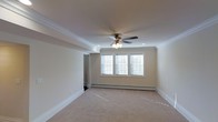 This single-floor space offers a clean and classic interior design with thoughtful architectural details throughout. The main room is generously proportioned, finished with soft, neutral-toned wall-to-wall carpeting that adds warmth and comfort underfoot. Crown molding lines the perimeter of the ceiling, lending an elegant and polished character to the space.

Overhead, a traditional ceiling fan with an integrated light fixture serves as a functional centerpiece, complemented by recessed lighting that provides even, ambient illumination throughout the room. The ceiling itself features a subtle stepped or tray detail, adding visual depth and dimension.

Natural light fills the room through a set of triple windows fitted with white horizontal blinds, allowing for flexible light control while maintaining a bright and airy atmosphere. A baseboard heating system runs along the exterior wall beneath the windows, ensuring efficient climate control.

The neutral color palette — soft beige walls paired with crisp white trim and molding — creates a versatile backdrop suitable for a variety of uses and furnishing styles. An open doorway visible along one wall suggests easy connectivity to adjacent areas. Overall, the space presents a well-maintained, move-in-ready interior with timeless finishes and a welcoming ambiance.