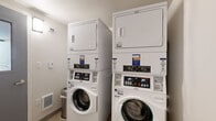 This shared laundry room is a practical, efficiently organized space designed to serve multiple users. The room houses several stacked washer and dryer units — each pair vertically stacked to maximize the use of available floor space. The machines are modern, front-loading washers paired with top-mounted dryers, and are operated through a coin meter system that also supports a mobile app for added convenience, allowing users to start machines and request service digitally.

The room features neutral white walls with a textured ceiling and light-toned flooring, creating a clean and functional atmosphere. A wall-mounted light fixture provides adequate illumination, while a low-profile floor vent ensures basic climate control. A small trash bin is conveniently placed near the machines for users' use.

Access to the room is through a solid gray door with a narrow window panel, allowing visibility into the space from outside. Instructional signage is posted on the walls, providing usage directions in multiple languages. Overall, the space is straightforward and utilitarian, prioritizing functionality and ease of use for shared laundry needs.