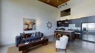 This single-floor space makes a striking first impression with its dramatic, soaring ceilings clad in warm wood planking, creating an airy yet intimate atmosphere. The open-concept layout seamlessly connects the living area to the kitchen, making the space feel expansive and well-integrated. The living area is anchored by a tufted charcoal Chesterfield sofa paired with a rustic wood coffee table and a classic accent chair, all resting on a neutral area rug over polished concrete floors. A bold, colorful longhorn painting adds a spirited focal point to the white walls, complemented by a decorative star wall ornament that lends artistic character. Track lighting and pendant lights work in tandem to provide both ambient and task illumination throughout. The kitchen features dark cabinetry contrasted by a clean white subway tile backsplash and light stone countertops, giving it a refined yet approachable look. A breakfast bar with wooden stools creates a casual dining area that flows naturally into the living space. The consistent use of warm tones, mixed textures, and carefully chosen décor gives this space a cohesive, urban-rustic personality that is both functional and visually engaging.
