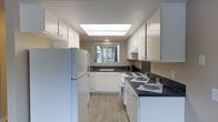 The kitchen is thoughtfully designed to maximize both functionality and aesthetics. White cabinetry lines the walls from floor to ceiling, offering generous storage space throughout. Dark countertops provide a striking contrast against the white cabinets and create a clean, modern look. The layout follows a U-shaped configuration, optimizing workflow and counter space for cooking and meal preparation. A skylight overhead floods the kitchen with natural light, complementing the window above the sink that looks out to the exterior. The light hardwood-style flooring adds warmth and continuity to the space. Full-size white appliances are included, featuring a refrigerator, an electric range with coil burners, and a range hood above the stove. A double basin sink is positioned beneath the window, providing a pleasant view while completing daily tasks. The overall design balances practicality with a bright, welcoming ambiance, making this kitchen both an efficient workspace and an inviting area of the home.