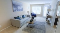 This single-floor space showcases a well-appointed living area designed with a clean, contemporary aesthetic. The room is finished with wall-to-wall carpeting in a warm neutral tone, complementing the crisp, light-colored walls that create an airy atmosphere throughout. Natural light flows generously into the space through large windows dressed with white shutters, with an additional glazed door providing further brightness and potential outdoor access.

The living area is thoughtfully arranged around a light gray sofa adorned with coordinating blue and green accent pillows, paired with a bold navy blue armchair that adds a pop of color. A low-profile rectangular coffee table with a chevron wood top and black metal frame serves as a stylish focal point, set atop a textured blue-gray area rug. Decorative ceramic vases in earthy tones add character to the tabletop. A large-scale cityscape artwork anchors one wall, lending a sophisticated, urban touch to the overall design. A floor lamp provides ambient lighting, contributing to the warm, inviting ambiance of the space.