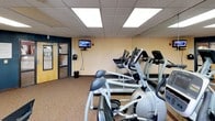 Located at 160 Sinclair St in Reno, Nevada, this fitness center offers a well-equipped, single-floor exercise facility designed to support a range of workout routines. The space is divided into distinct functional zones, with a dedicated cardio area housing multiple treadmills, elliptical trainers, and a stair climber, all positioned along mirrored walls that enhance the sense of space and allow users to monitor their form. A wall-mounted television provides entertainment during cardio sessions. The strength training area features a multi-station cable and weight machine alongside additional equipment such as an exercise bike and stability ball, offering versatility for both resistance and flexibility training. The flooring transitions between durable carpet and rubberized surfaces suited to the activity in each zone. Bold navy blue accent walls contrast with neutral cream tones, creating an energizing atmosphere. Overhead, a drop ceiling with recessed panel lighting ensures consistent, bright illumination throughout the room. Natural light filters in through multiple windows overlooking surrounding greenery, adding a refreshing quality to the environment. A wall-mounted HVAC unit provides climate control, keeping the space comfortable year-round. The facility is accessible via a glass-paneled entry door and is thoughtfully organized to maximize usable floor space for an effective workout experience.