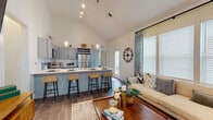 This single-floor residence is defined by its striking vaulted ceilings, which lend an airy, expansive quality to the open-concept main living area. The kitchen is a functional centerpiece, outfitted with grey-blue shaker cabinetry, white quartz countertops, stainless steel appliances—including a refrigerator, range, and microwave—and a peninsula with seating for multiple guests. Pendant lighting and a tile backsplash add refined detail. The kitchen flows seamlessly into the living area, which features wide-plank wood-style flooring, large windows with decorative drapery, and abundant natural light throughout.

The bedroom is well-proportioned, offering multiple windows, a ceiling fan, and direct access to a walk-in closet with built-in shelving. The primary bathroom is spacious, featuring a dual-sink vanity with grey cabinetry and quartz countertops, a glass-enclosed walk-in shower, and wood-style flooring. A secondary bathroom includes a single vanity and convenient access to an in-unit laundry area with full-size washer and dryer. Throughout the residence, a neutral color palette and consistent finishes create a cohesive, modern aesthetic.