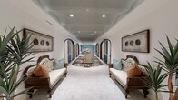Located at 98 South River Drive in Tempe, Arizona, this single-floor community space is a masterclass in refined interior design. The grand entrance corridor immediately sets the tone, featuring a dramatic series of dark wood-trimmed arches flanked by navy blue built-in shelving and cabinetry, centered by a vibrant, artisan area rug and a sculptural lounge chair — a hallway that doubles as a gallery-like experience leading to the branded reception area of The Alyssa.

The living room is a showstopper, boasting soaring double-height ceilings lined with an abundance of oversized windows that flood the space with abundant natural light. Eclectic furnishings — including a rich plum sofa, warm leather chairs, and cowhide rugs — are arranged to encourage both intimate conversation and social gathering, while a large-screen TV is integrated into teal cabinetry along one wall.

The dining area is anchored by a striking deep-teal curved bar with a marble countertop and decorative lattice detailing overhead, providing a sophisticated focal point. Adjacent lounge seating areas feature curated artwork and warm amber upholstery. Entry vestibules showcase polished marble-look tile flooring and herringbone brick accents, with warm gold and taupe finishes completing a cohesive, luxurious aesthetic throughout.