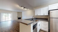 This single-floor residence offers a thoughtfully designed open-concept layout with dark hardwood-style flooring running consistently throughout. The kitchen serves as a central hub, featuring white shaker-style cabinetry with brushed nickel hardware, dark grey countertops, and a mosaic tile backsplash. A peninsula with an integrated sink and built-in dishwasher provides additional workspace and naturally separates the kitchen from the living area. Stainless steel appliances, including a refrigerator, range, and over-the-range microwave, complete the kitchen. Track lighting illuminates both the kitchen and the adjacent living area.

The spacious living room is anchored by a corner stone fireplace with a structured mantel, adding warmth and visual interest to the space. A sliding glass door connects the living area to an outdoor space, allowing natural light to flow in. The bedrooms offer generous proportions, neutral-toned walls, and sliding closet doors. One bedroom also features a sliding glass door providing additional outdoor access.

The bathroom is finished with a full tub-and-shower combination enclosed by a sleek sliding glass door with brushed nickel fixtures, complementing the clean, contemporary aesthetic maintained throughout the residence.