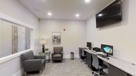This single-floor business center offers a well-organized and functional environment suited for both focused work and casual meetings. The space is finished with neutral gray walls and recessed lighting that create a bright, professional atmosphere throughout. A built-in granite-topped workstation along one wall accommodates multiple desktop computers and a printer, providing residents or visitors with the tools needed for everyday tasks. Ergonomic rolling chairs are positioned at each station for comfort during extended use. A wall-mounted flat-screen display is installed above the workstation area, adding a modern touch. On the opposite side of the room, a relaxed seating arrangement features upholstered gray armchairs paired with a sleek glass side table and a decorative lamp, offering a comfortable spot for informal conversations or reading. A framed cityscape artwork adds a refined accent to the otherwise minimalist décor. Carpeted flooring in a subtle pattern contributes to the room's understated elegance while providing acoustic comfort. Natural light filters in through a window on one side, complementing the recessed ceiling lights to keep the space well-illuminated and inviting.