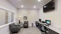 This single-floor business center offers a well-organized and functional environment suited for both focused work and casual meetings. The space is finished with neutral gray walls and recessed lighting that create a bright, professional atmosphere throughout. A built-in granite-topped workstation along one wall accommodates multiple desktop computers and a printer, providing residents or visitors with the tools needed for everyday tasks. Ergonomic rolling chairs are positioned at each station for comfort during extended use. A wall-mounted flat-screen display is installed above the workstation area, adding a modern touch. On the opposite side of the room, a relaxed seating arrangement features upholstered gray armchairs paired with a sleek glass side table and a decorative lamp, offering a comfortable spot for informal conversations or reading. A framed cityscape artwork adds a refined accent to the otherwise minimalist décor. Carpeted flooring in a subtle pattern contributes to the room's understated elegance while providing acoustic comfort. Natural light filters in through a window on one side, complementing the recessed ceiling lights to keep the space well-illuminated and inviting.